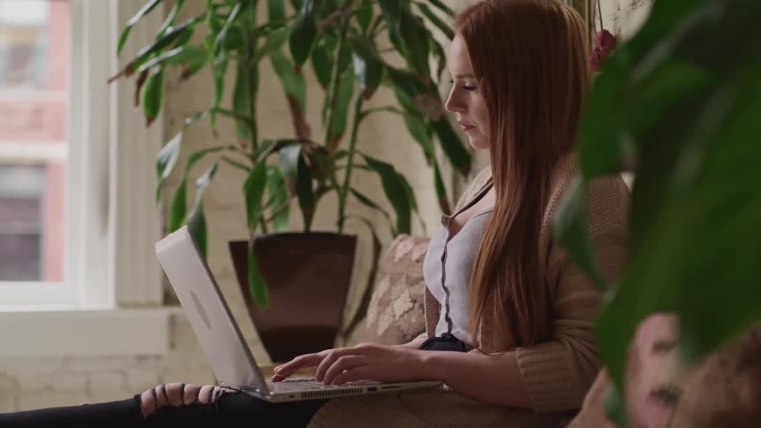 A woman types on a laptop while sitting on a sofa in an apartment
