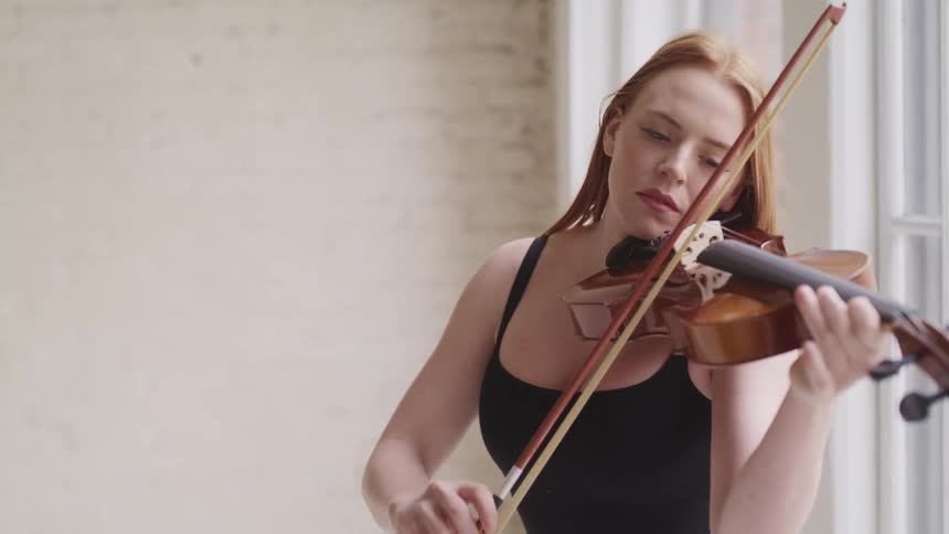 Woman with fair skin red hair and a black dress holds violin against her neck and plays by moving a bow