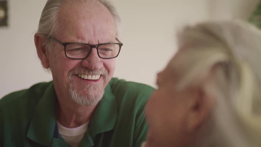 Elderly man with a goatee beard a green polo shirt and glasses looks into elderly womans eyes and laugh