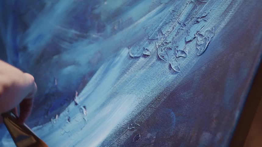 Artist paints lighter blue across darker blues on canvas using large brush