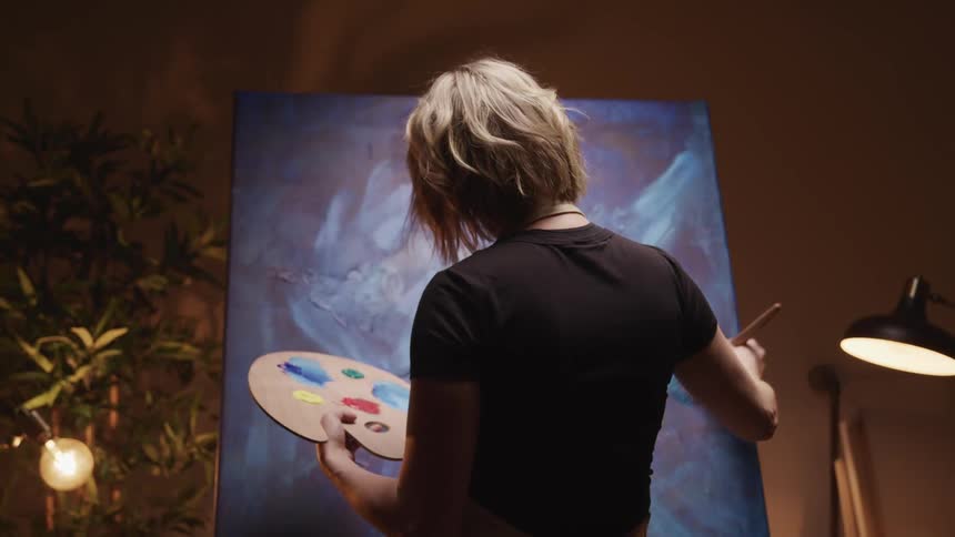 Artist holds palette and uses large brush to work on painting containing a lot of blues in studio lit by lamps