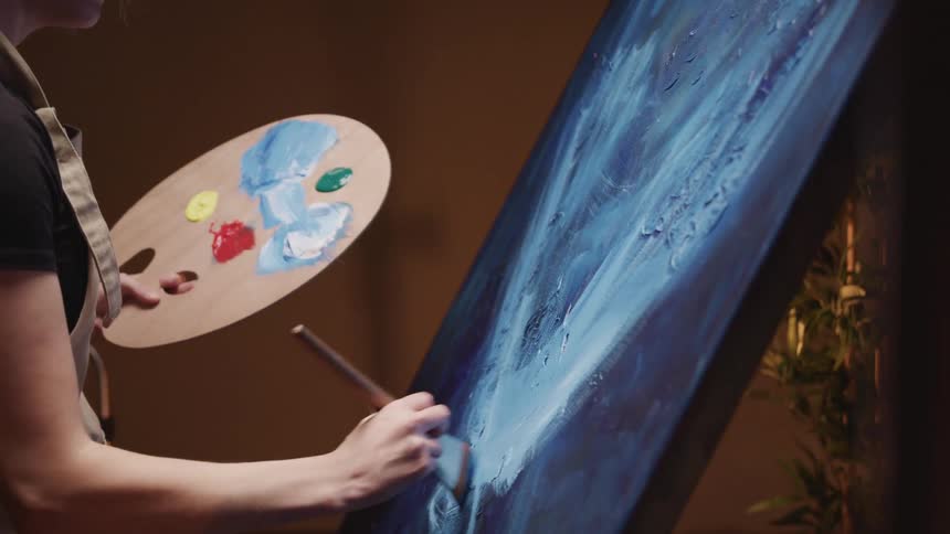 An artist holds a palette in one hand while she paints white over blue on a canvas