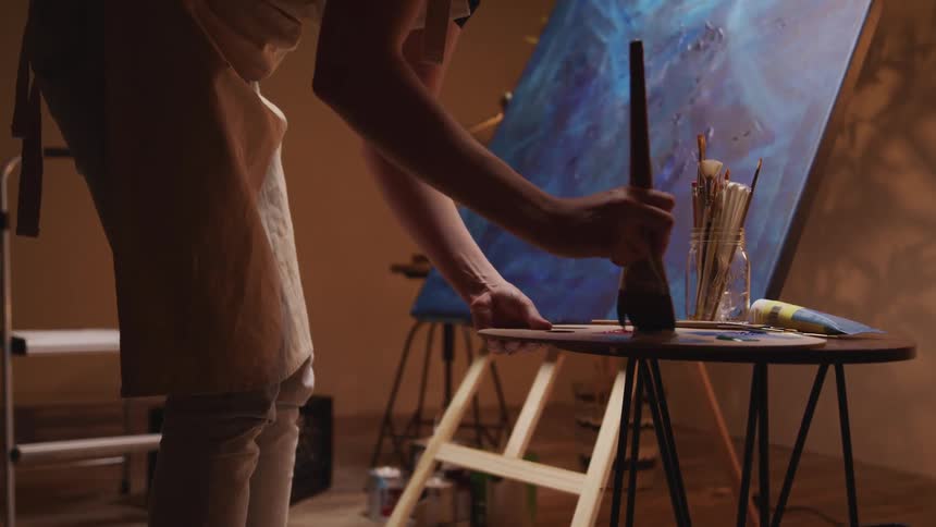 Artist dabs a paintbrush on a paint palette as artwork sits nearby on an easel