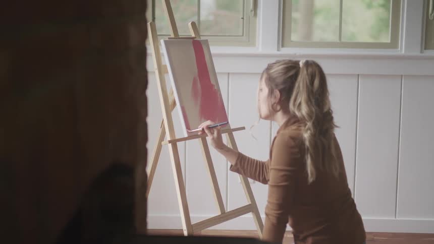 A woman sits on the floor and paints on a canvas sitting on an easel