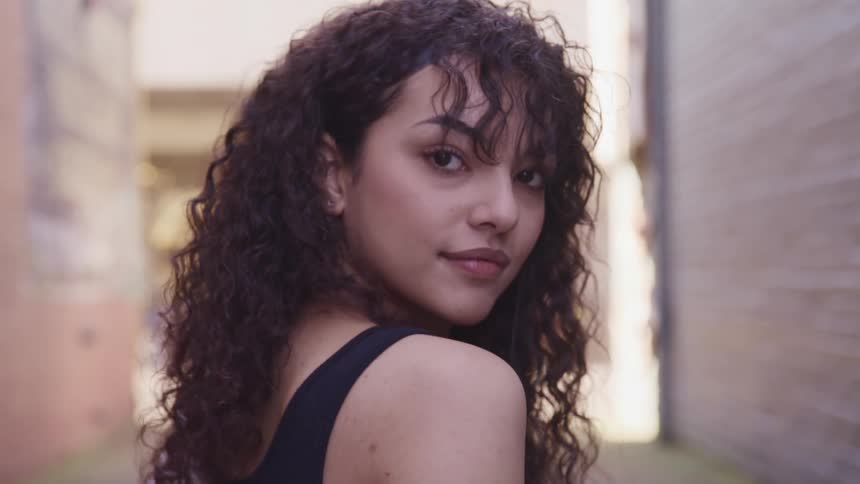 A young woman standing in an alleyway turns her head to look over her shoulder and into the camera