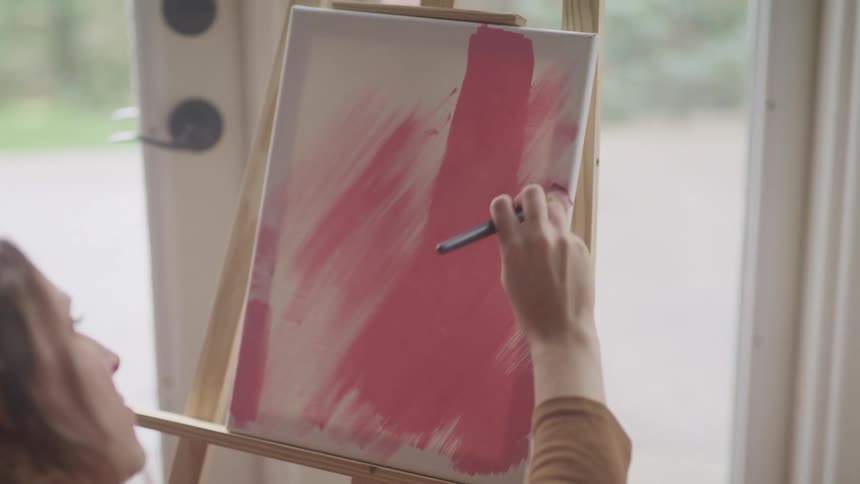 A woman paints pink strokes on a small canvas as she sits in front of an easel near a window a sunny day