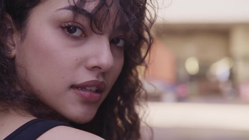A young woman stands outdoors in a city and looks back over shoulder into the camera