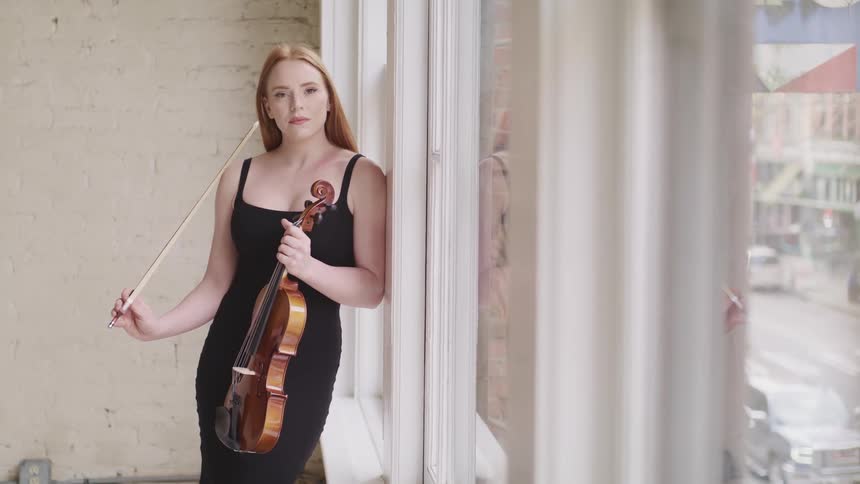 Woman with fair skin red hair and a black dress holds a violin and leans against a wall