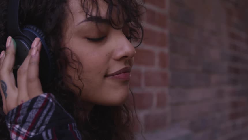 A woman standing listening to music on headphones with a brick wall in the background