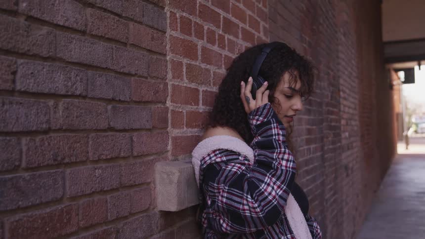 Woman with curly hair leans against a brick wall and listens to music with black headphones in an alley