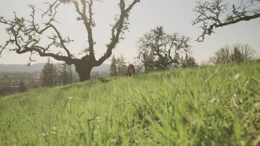 A person sitting on a grassy field with trees the sky and clouds in the background