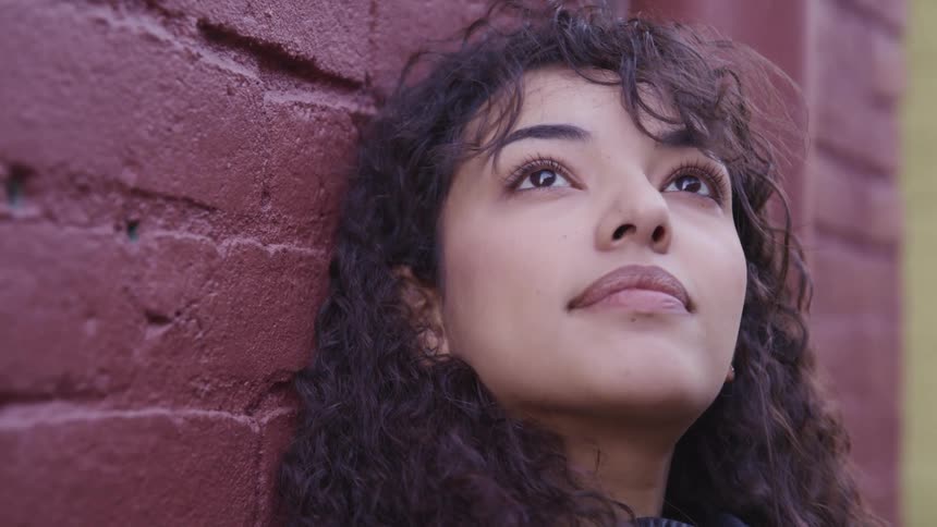 A young woman gazes up at the sky while leaning against a building