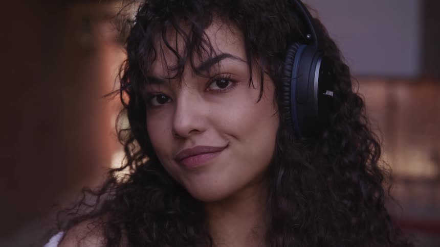 Woman with curly hair and black headphones faces the camera with eye contact and smile in an alley