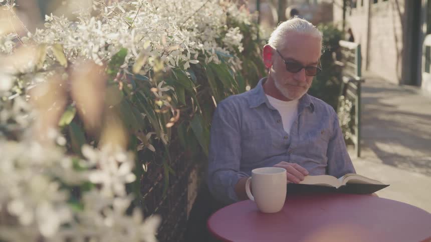 A man sitting at a cafe table next to a flowercovered wall reads a book next to a coffee cup