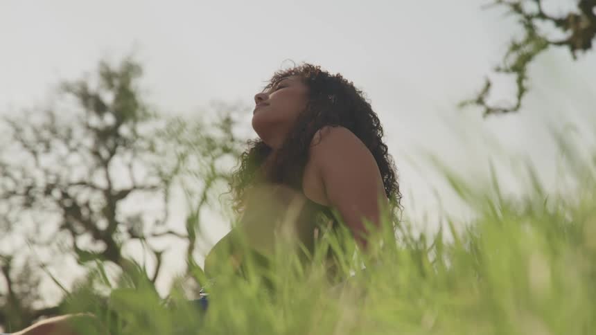 A young woman rests her eyes while sitting in tall grass under a pale sky