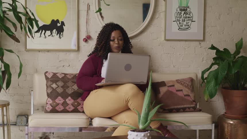 A black woman sitting on a sofa looks up from her laptop to smile into the camera