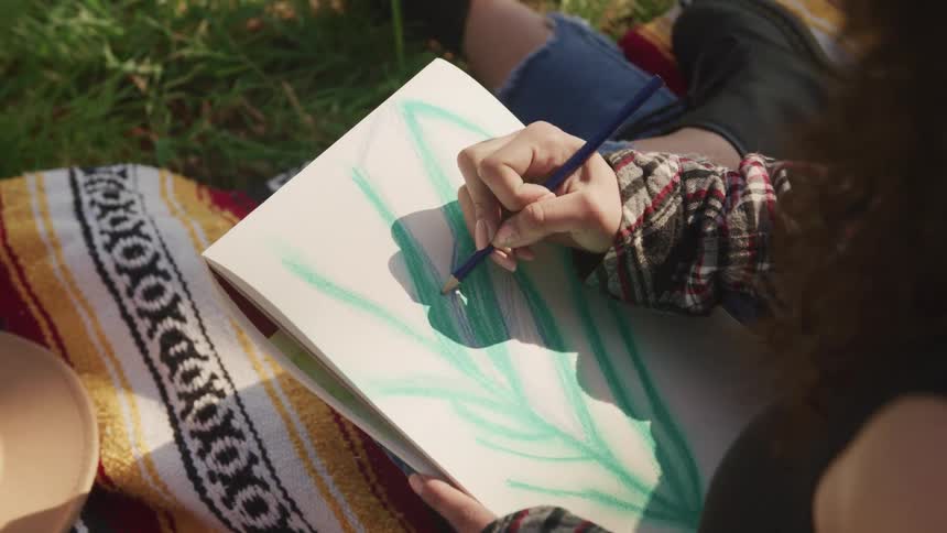 A woman with a pad drawing while sitting on a blanket on grass
