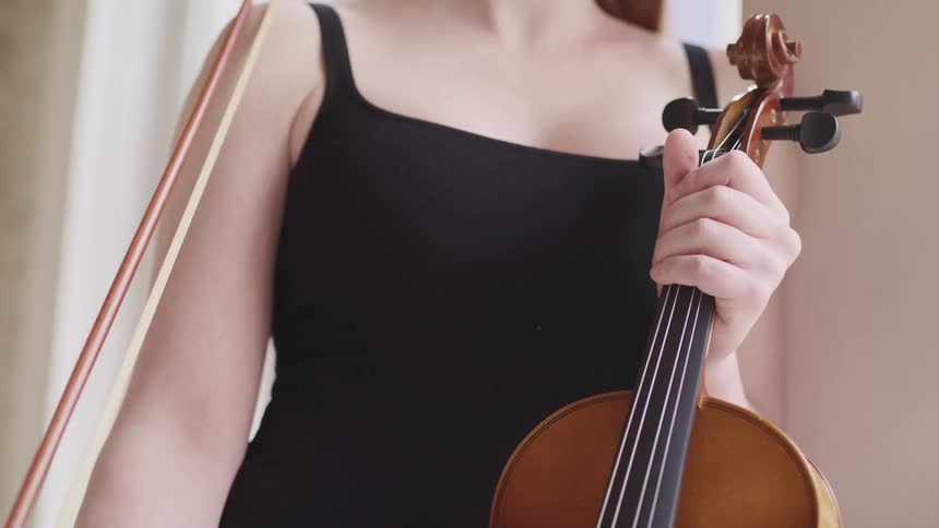 A woman stands near a window while holding a violin and bow