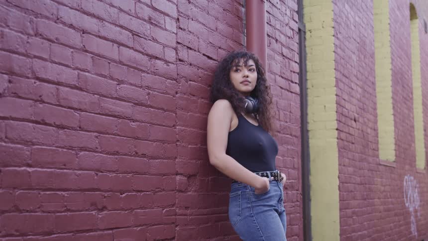 Woman with curly hair and headphones leans against red brick wall and turns her head to the right