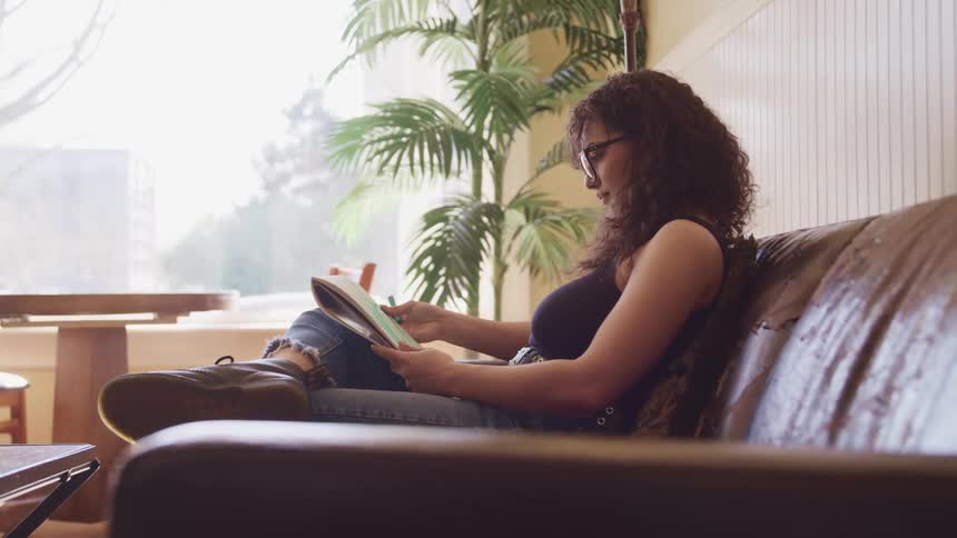 Female uses finger to blend colors on drawing she is making while sitting on the sofa in a living room apartment and then begins to draw again using green artist chalk