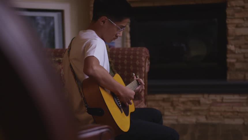Teenage boy with a white t shirt and glasses sits on a red sofa and plays a guitar by pulling the strings with his fingers next to the fireplace in the living room