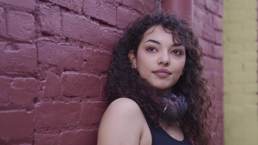 A woman in a tank top and scarf with long curly hair leans against a red painted brick wall
