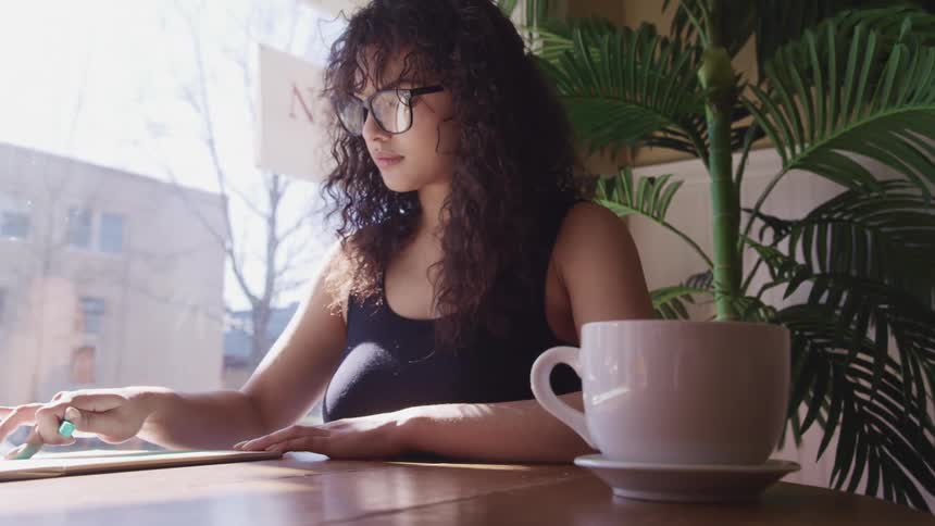 A woman sits at a cafe table with a coffee mug and folding a piece of paper carefully