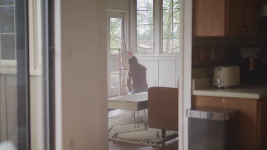 A woman sits on a stool in the living room of a house while painting on a small canvas on an easel