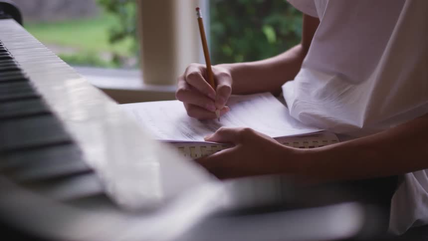 A young female sits at a piano and writes musical notes in a notebook