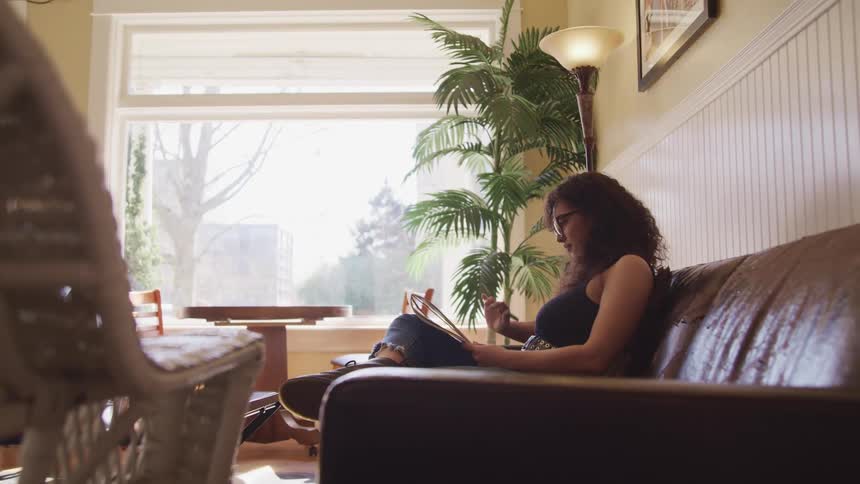A young woman reads from a notepad as she sits on a leather sofa in the living room of a home
