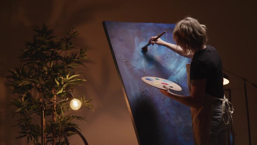 A person with a apron a paint brush and painting pallet painting on a canvas with a lamp and a plant in the background