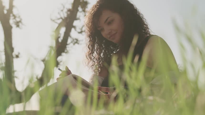 Woman contemplates sketch pad and brushes back her hair while standing among grass near tree