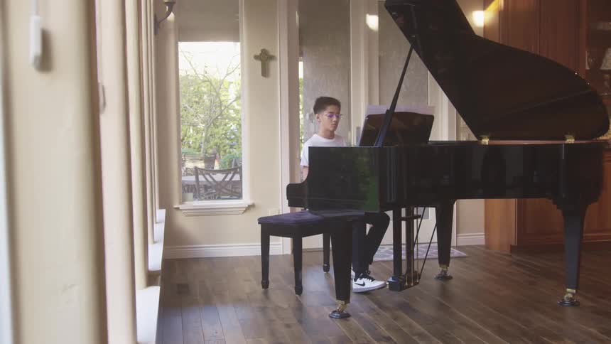 Teenage boy with a white t shirt and glasses sitting on a piano stool plays a grand piano