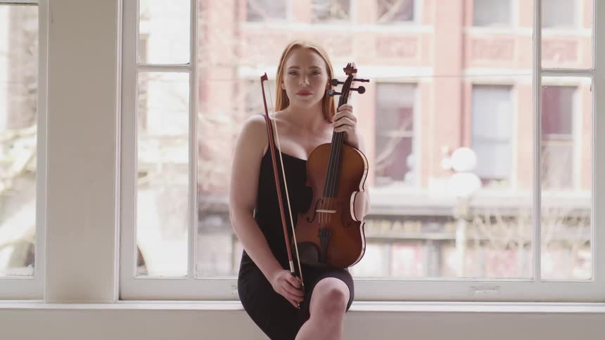 Woman with red hair and a black dress holds a violin and sits on windowsill