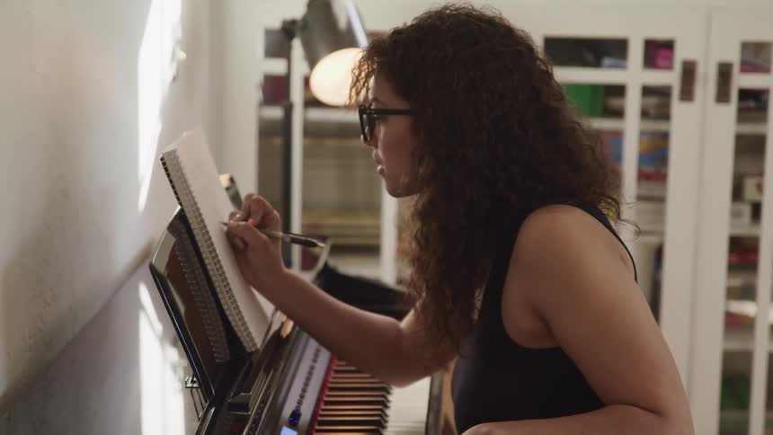 Woman with curly hair and glasses plays a piano and takes notes in a room