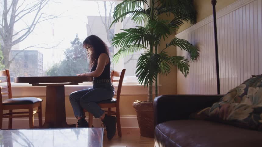 A young woman sits at a table in front of window and uses a highlighter to highlight items on a sheet of paper