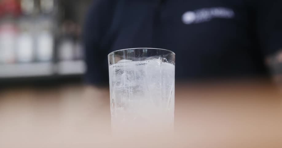 A tattooed bartender pours a bottle of liquid into a cup of ice and mixes it with a spoon