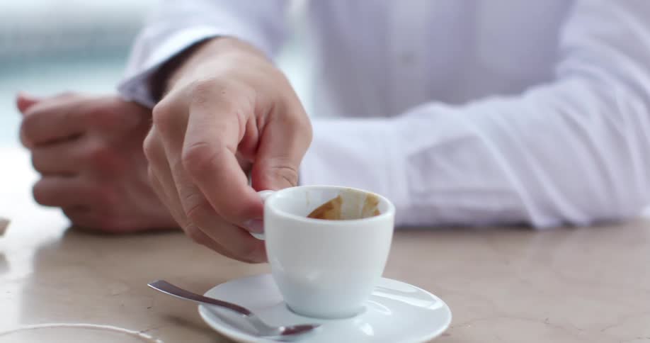 Man with a white shirt sits at a table and drinks a cup of espresso