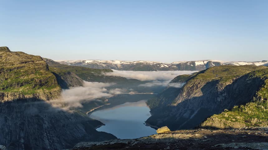Mist forms near the hills and river as snow covered mountains sit in the distance under a blue sky