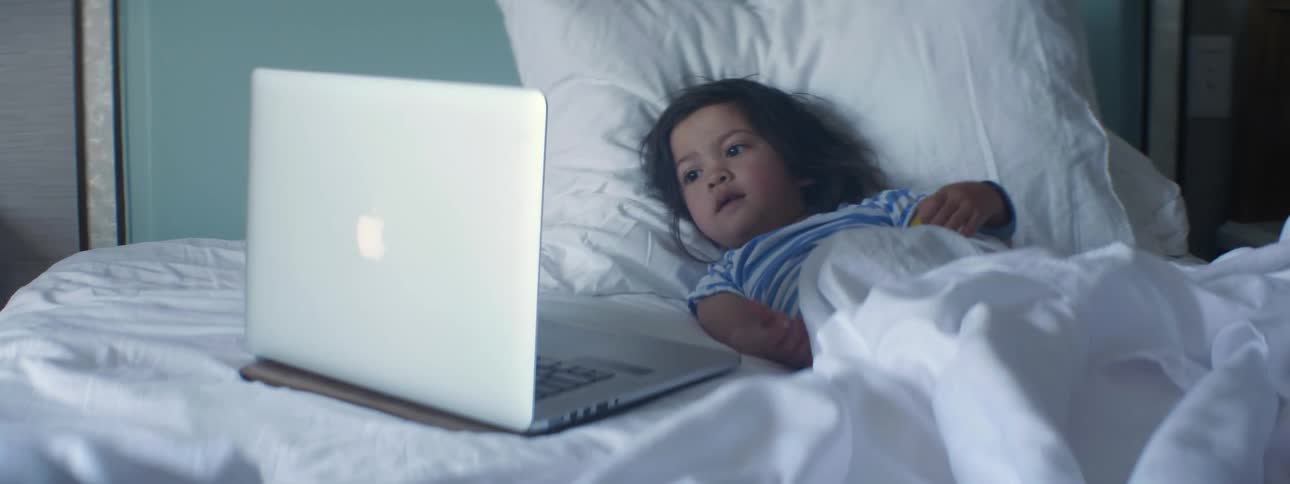 A little girl fiddles with a toy while lying in bed looking at the screen of a laptop lying next to her