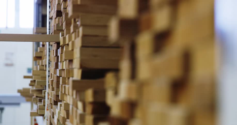 A person adds a wooden plank to a stack of wood planks on a shelf in a warehouse