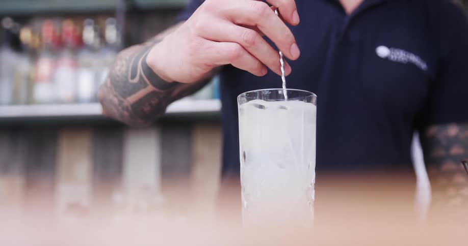 Bartender with tattoo sleeves stirs pale drink in tall glass