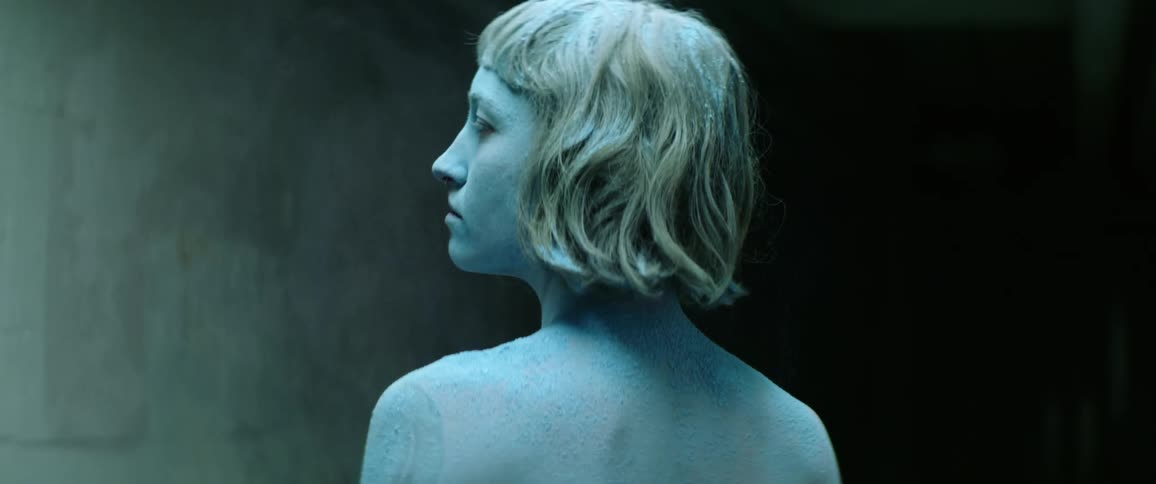 A naked young woman covered in blue powder stands in a dark hallway while slowly turning her head to look into the camera