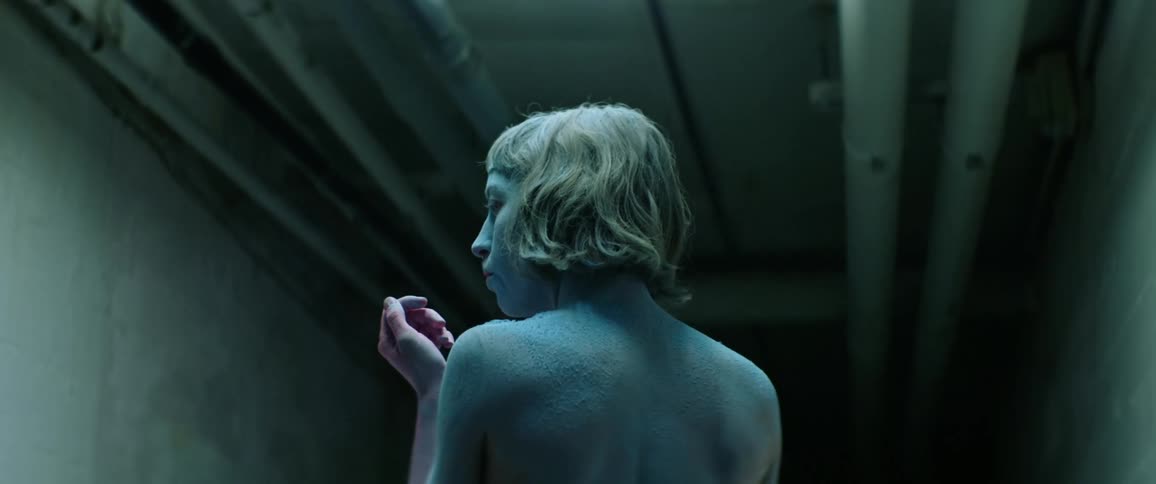A woman covered in blue powder blows pink powder off of her hand before turning to look down a dark corridor