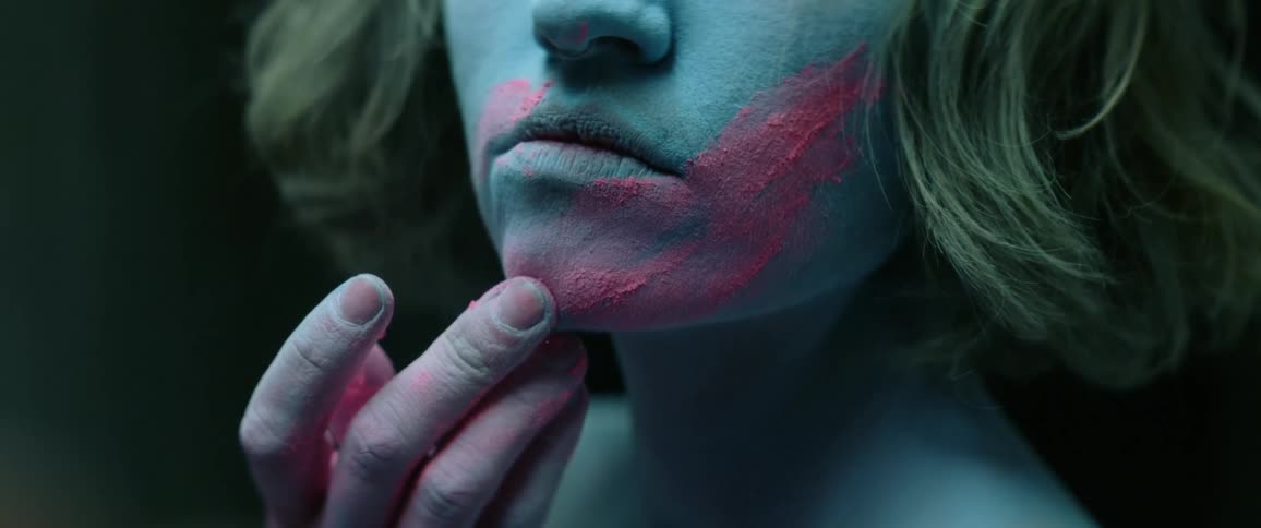 A naked woman covered in blue powder uses her fingers to smear pink powder over her lower face