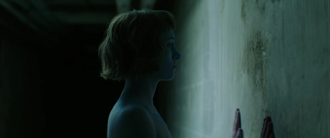 A naked young woman faces and slides her hands down a concrete wall in a dimly lit corridor