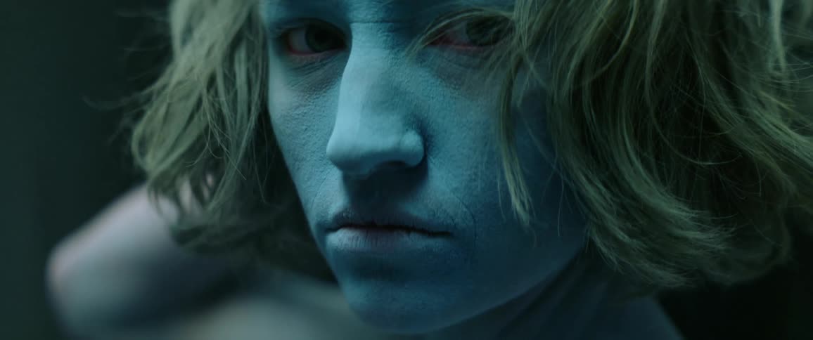 A young woman whose face is covered in blue powder turns her head to look into the camera with a stern expression