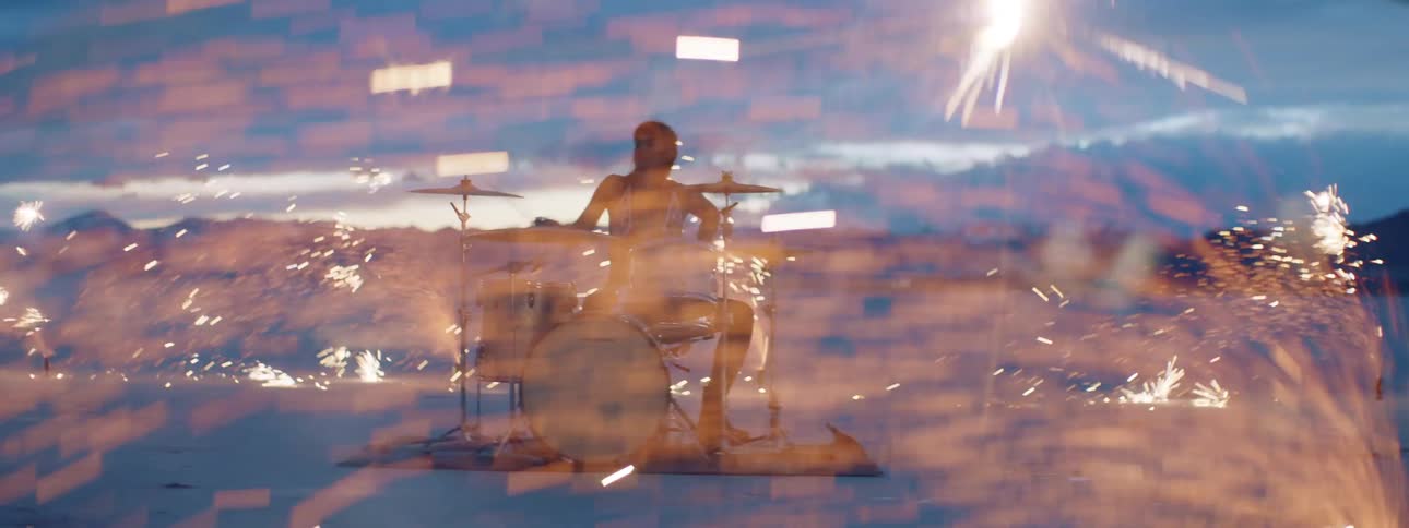 A person sitting down playing drums in the desert surrounded by fireworks with the sky in the background