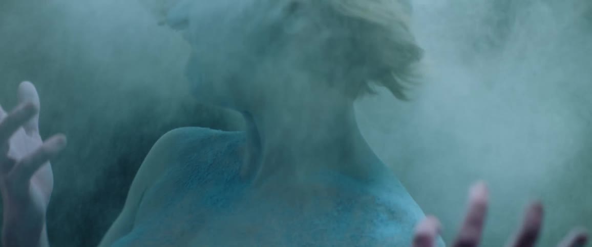 Young woman covered in colored powder shakes her hair violently in a dimly lit room