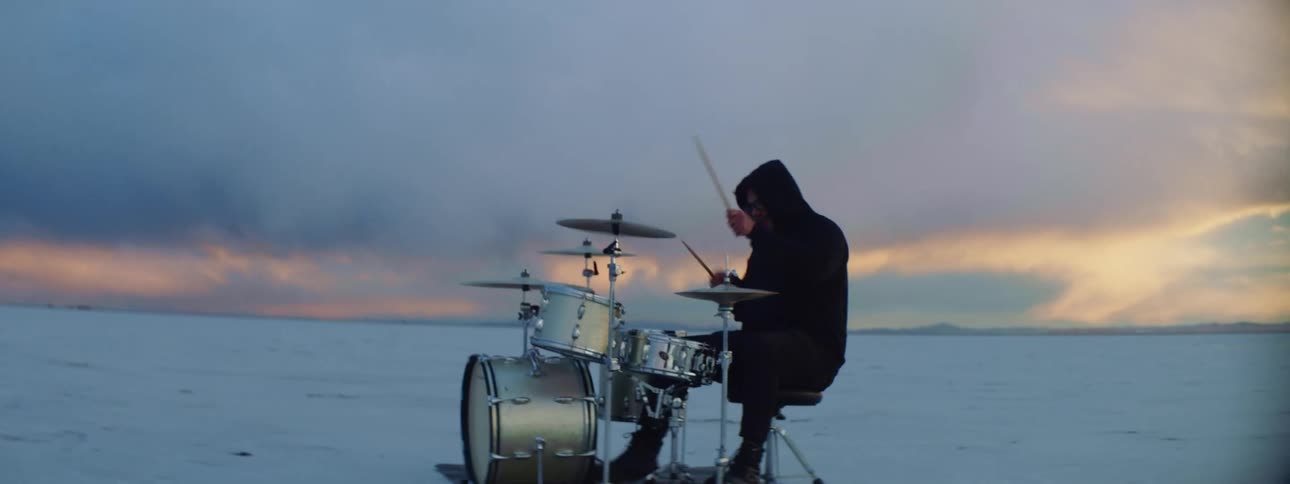 A man with a hoodie sitting playing drums in the desert with the sky in the background